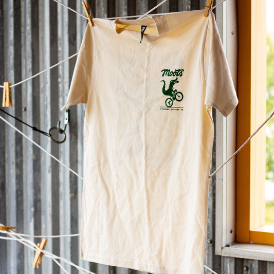 A cream-colored short-sleeve t-shirt with a green graphic print hanging on a clothesline against a corrugated metal wall.