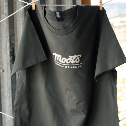 Pine green Moots Staple Tee hanging on a clothesline against a corrugated metal wall, with the Moots script logo and "Steamboat Springs, CO." printed in white on the chest.