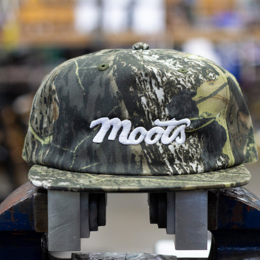 A camouflage baseball cap with 'Moots' embroidered in white script on the front, displayed in a blue bench vise against a blurred workshop background.