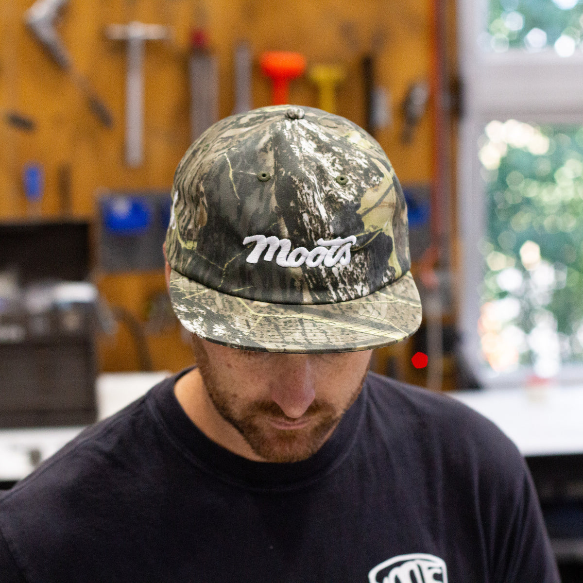 Close-up of a man wearing the Moots Camo Hat with the white embroidered Moots script logo, in the workshop.