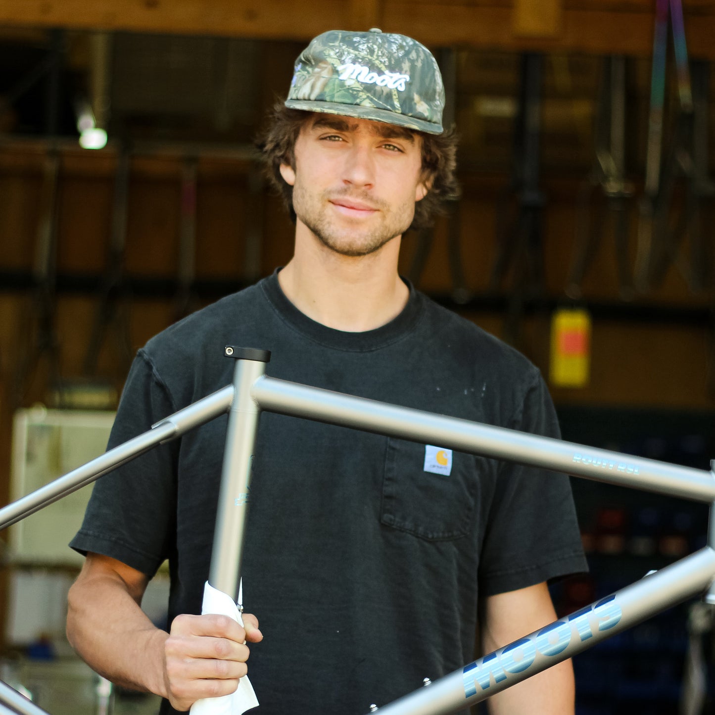 A builder wearing the Moots Camo Hat holds a titanium Moots bike frame in the workshop.