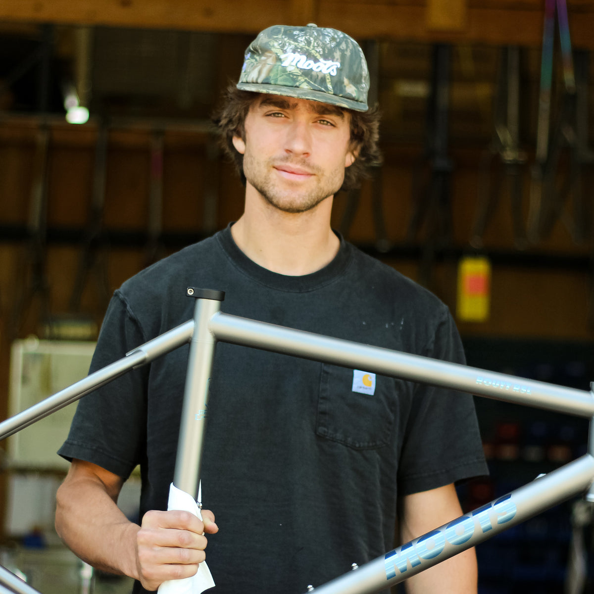A builder wearing the Moots Camo Hat holds a titanium Moots bike frame in the workshop.