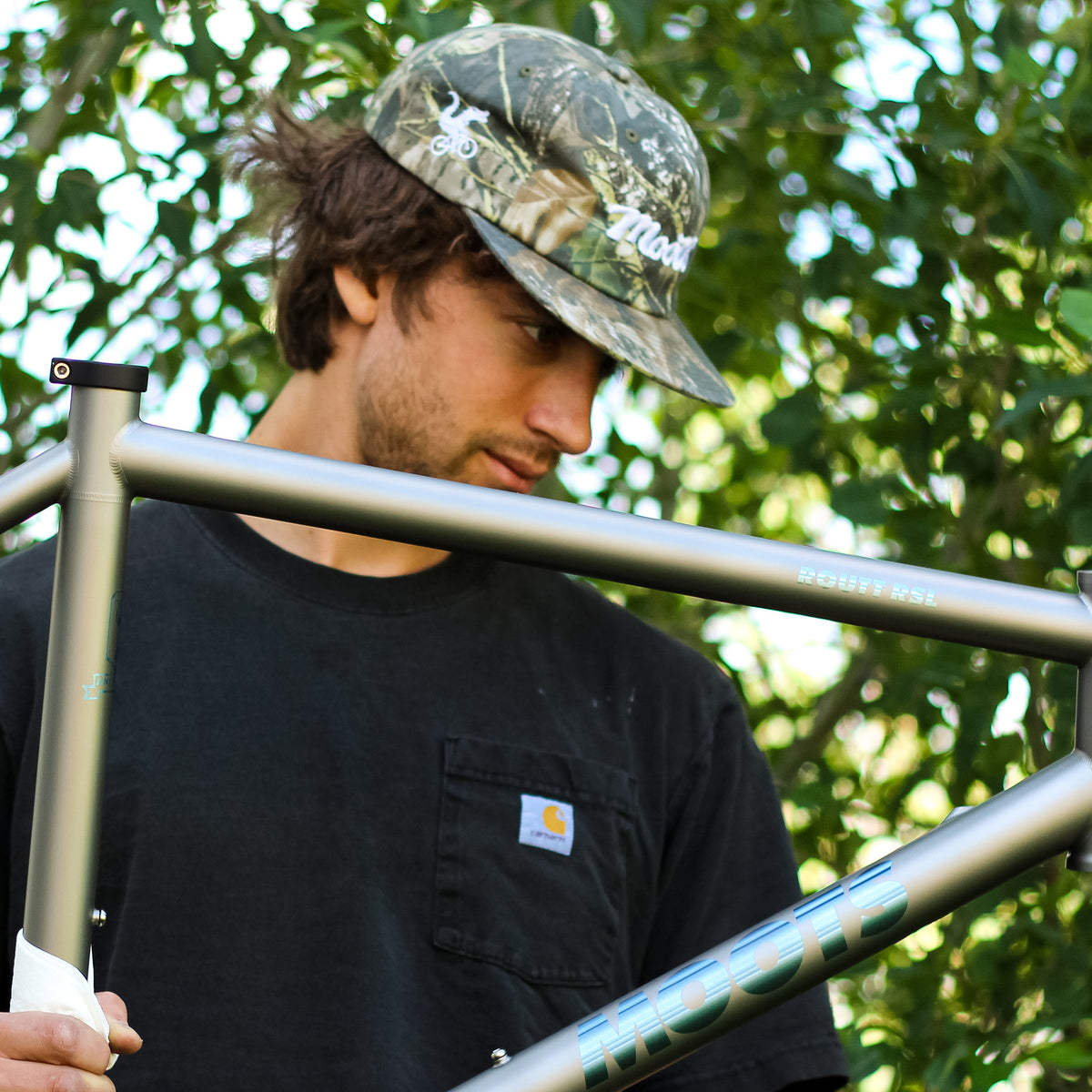 A man wearing the Moots Camo Hat holds a titanium Moots bike frame outdoors among green trees.